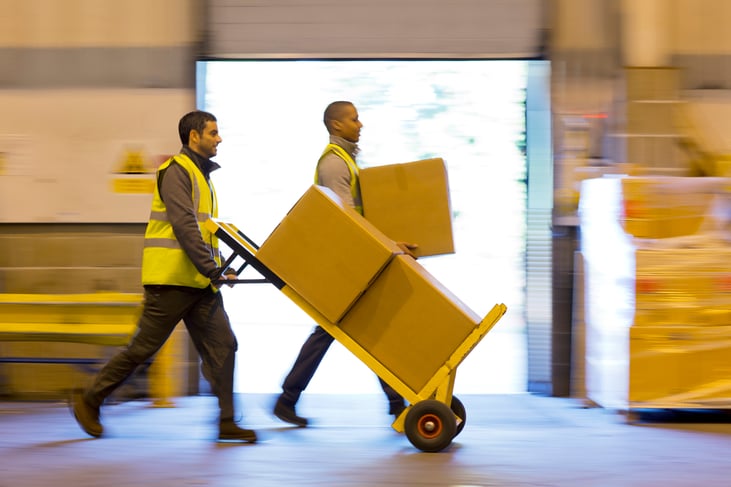 Two workers carting boxes in warehouse quickly, wearing high visibility vests