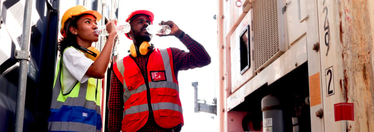 Two industrial workers engineers wearing safety vests and headgear drinking water from bottle during a break after working hard at logistic shipping cargo container yard.